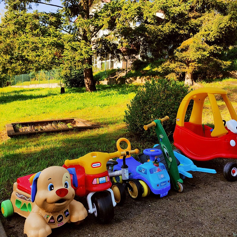 Kolorowe zabawki dla dzieci, w tym piesek na kółkach, jeździk i hulajnoga, ustawione na zewnątrz na tle trawnika i drzew.