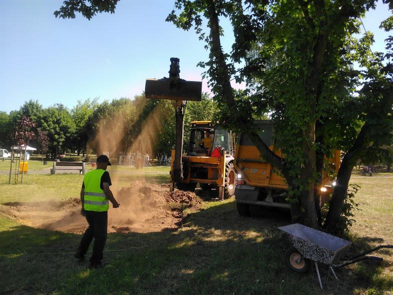 Operator koparki w kamizelce odblaskowej nadzoruje wykop ziemi w parku, pył unosi się w powietrzu, w tle widać przyczepę i drzewa.