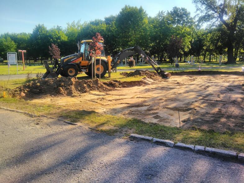 Żółta koparko-ładowarka Volvo podczas prac ziemnych na terenie parku, widoczne wykopane rowy i pryzma ziemi, w tle drzewa i elementy placu zabaw.