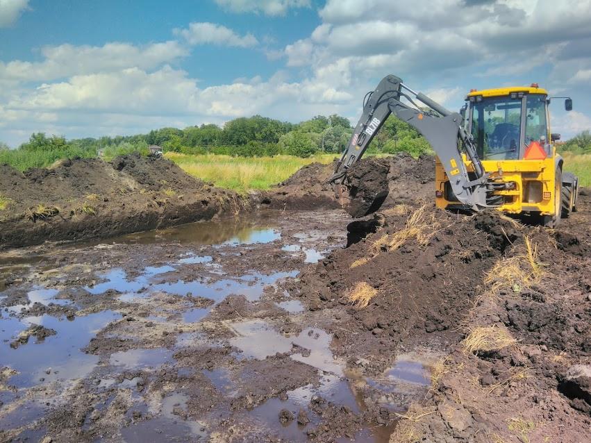Żółta koparko-ładowarka nabiera błotnistą ziemię z wykopu na tle zieleni i błękitnego nieba.