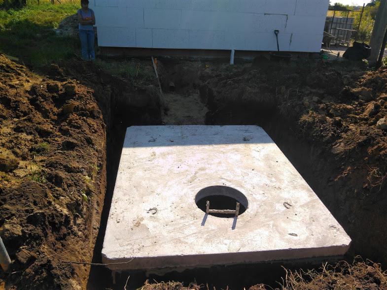 Betonowa płyta z otworem w wykopie, widoczna osoba w tle przy budynku z bloczków.
