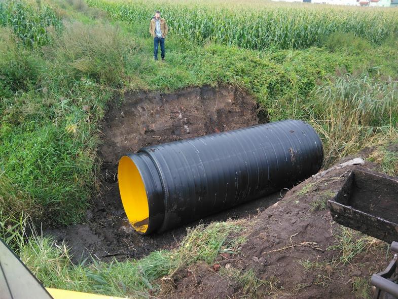 Czarna, karbowana rura z żółtym wnętrzem ułożona w wykopie ziemnym; w tle pole kukurydzy i postać mężczyzny.