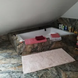 Biała wanna w obudowie z płytek imitujących kamień, różowy dywanik łazienkowy na podłodze z podobnych płytek, półka z ręcznikami i kosmetyki w łazience na poddaszu.