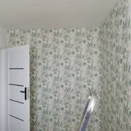 Ściana z tapetą w motywy tropikalne, widoczna biała ościeżnica drzwi z czarnymi poziomymi paskami i fragment drabiny aluminiowej.