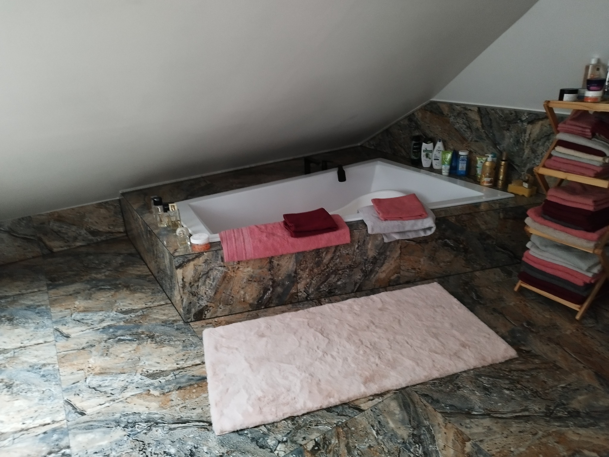 Biała wanna w obudowie z płytek imitujących kamień, różowy dywanik łazienkowy na podłodze z podobnych płytek, półka z ręcznikami i kosmetyki w łazience na poddaszu.