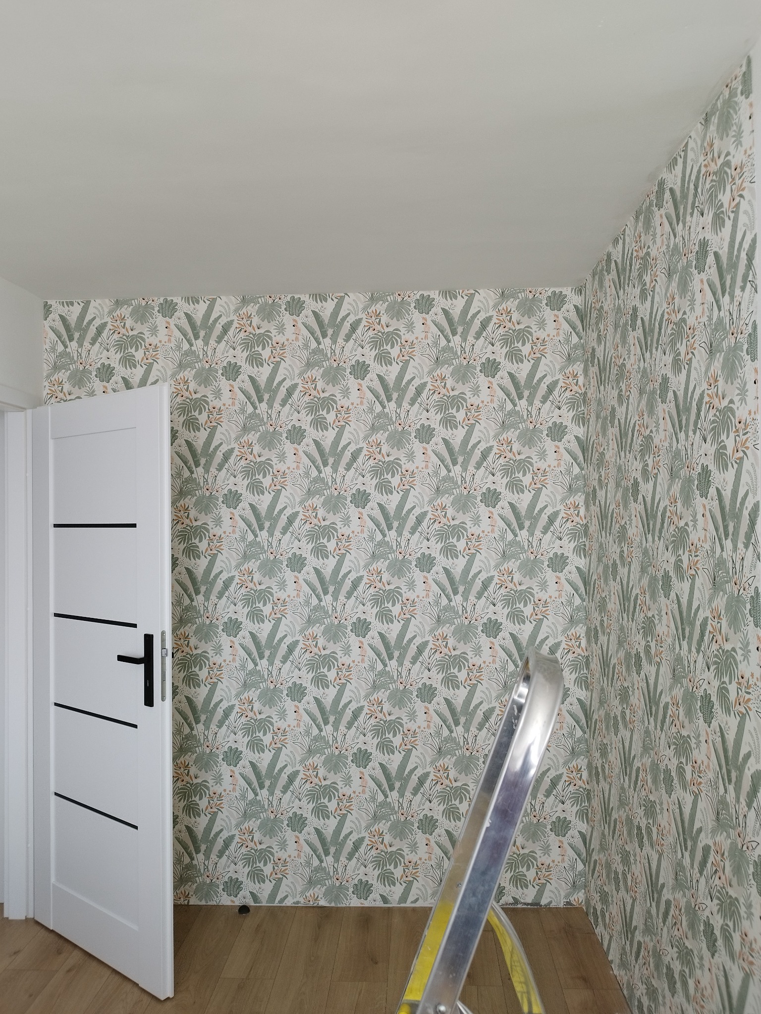 Ściana z tapetą w motywy tropikalne, widoczna biała ościeżnica drzwi z czarnymi poziomymi paskami i fragment drabiny aluminiowej.
