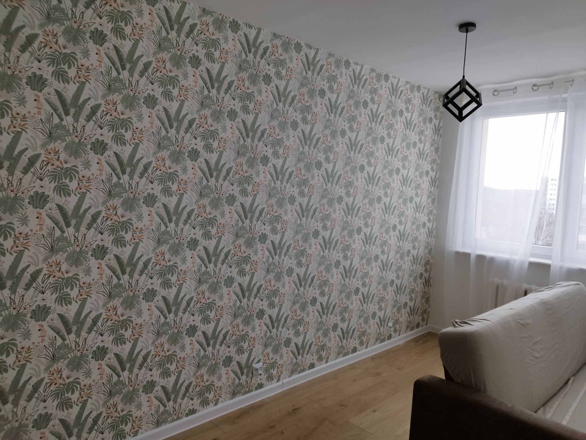 Ściana w pokoju pokryta tapetą z motywem dżungli, widoczna lampa wisząca o geometrycznym kształcie, fragment sofy i okno z białymi firankami.