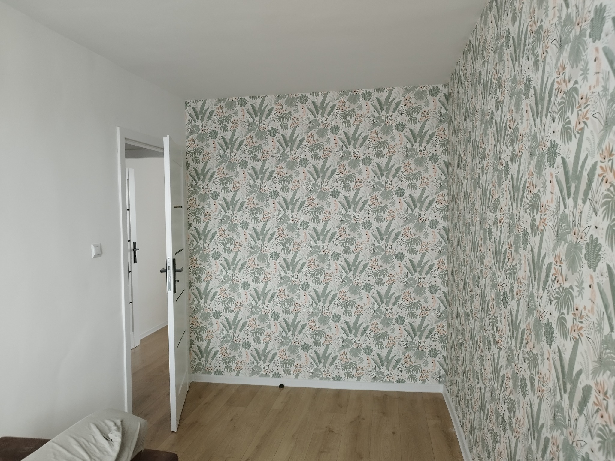 Pokój po remoncie z nową tapetą w tropikalne wzory na jednej ścianie i świeżo pomalowanymi na biało pozostałymi ścianami, widoczne drewniane panele podłogowe i otwarte białe drzwi.