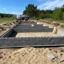 Fundamenty domu jednorodzinnego w trakcie budowy, bloczki betonowe ułożone w wykopie wypełnionym piaskiem, widoczne rury kanalizacyjne i zaparkowany bus budowlany w tle.