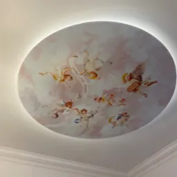 Okrągła dekoracja sufitowa z motywem aniołków na tle chmur, podświetlona od tyłu, widoczna z dołu, z detalami sztukaterii na styku ze ścianą.