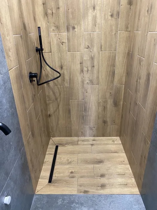 Kabina prysznicowa wyłożona płytkami imitującymi drewno, z czarną armaturą i odpływem liniowym.
