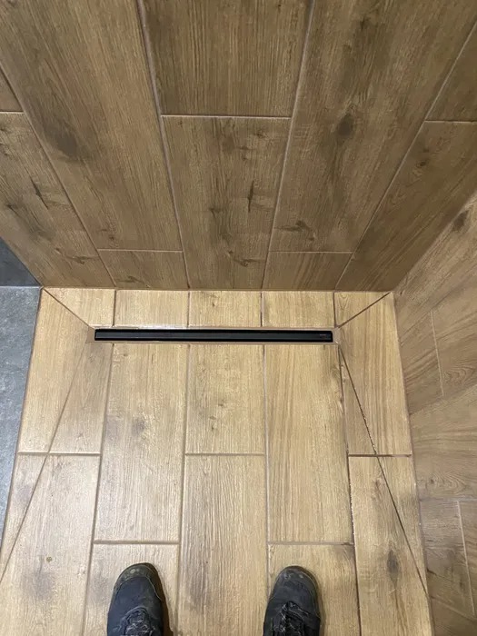 Wykończona kabina prysznicowa z odpływem liniowym i drewnopodobnymi płytkami na ścianach i podłodze, widoczne buty osoby robiącej zdjęcie.