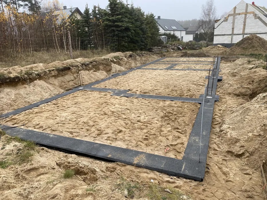 Przygotowany teren pod fundamenty domu jednorodzinnego z widocznymi czarnymi panelami izolacyjnymi i wystającymi prętami zbrojeniowymi.