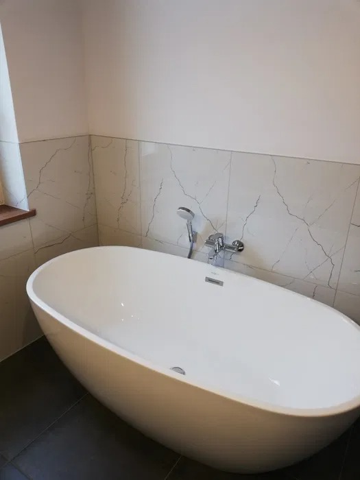 Nowoczesna, wolnostojąca wanna owalna z chromowaną baterią i słuchawką prysznicową na tle marmurowych płytek ściennych z delikatnym żyłkowaniem.