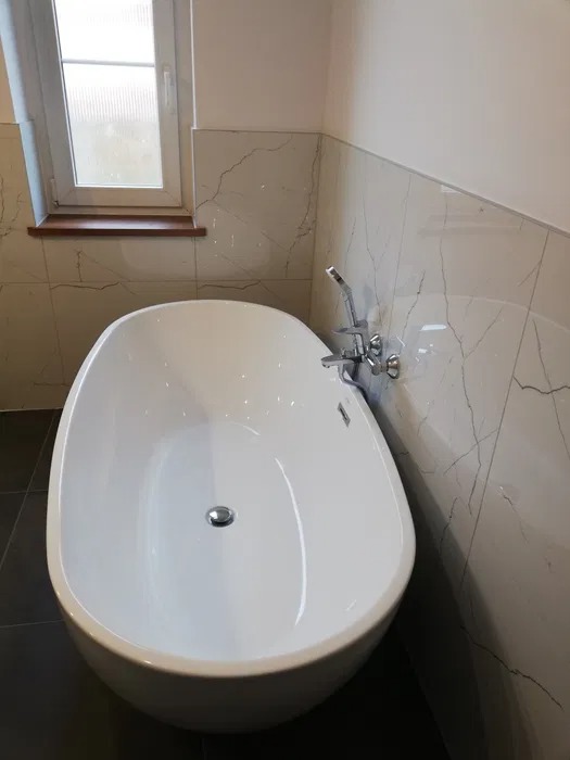 Wolnostojąca, owalna wanna w narożniku łazienki wyłożonej płytkami imitującymi marmur, widoczne okno z drewnianym parapetem i chromowana bateria.