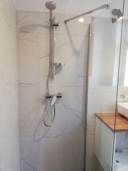 Nowoczesny prysznic walk-in z białą armaturą i płytkami imitującymi marmur z wyraźnym żyłowaniem, częściowo widoczna umywalka nablatowa i drewniany blat szafki.