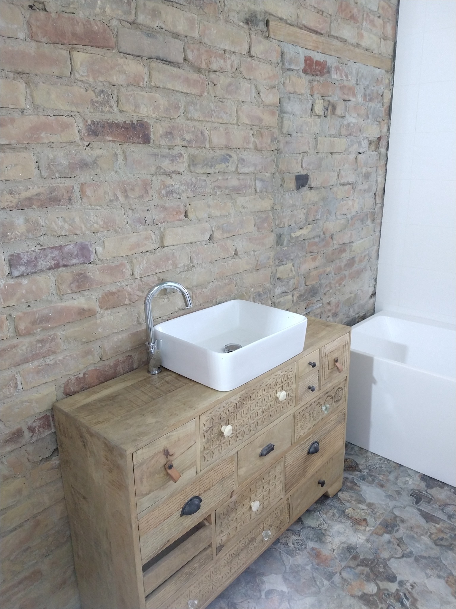 Biała, prostokątna umywalka nablatowa z chromowaną baterią na drewnianej komodzie z rzeźbionymi frontami szuflad, na tle ściany z cegły i fragmentu białej wanny.
