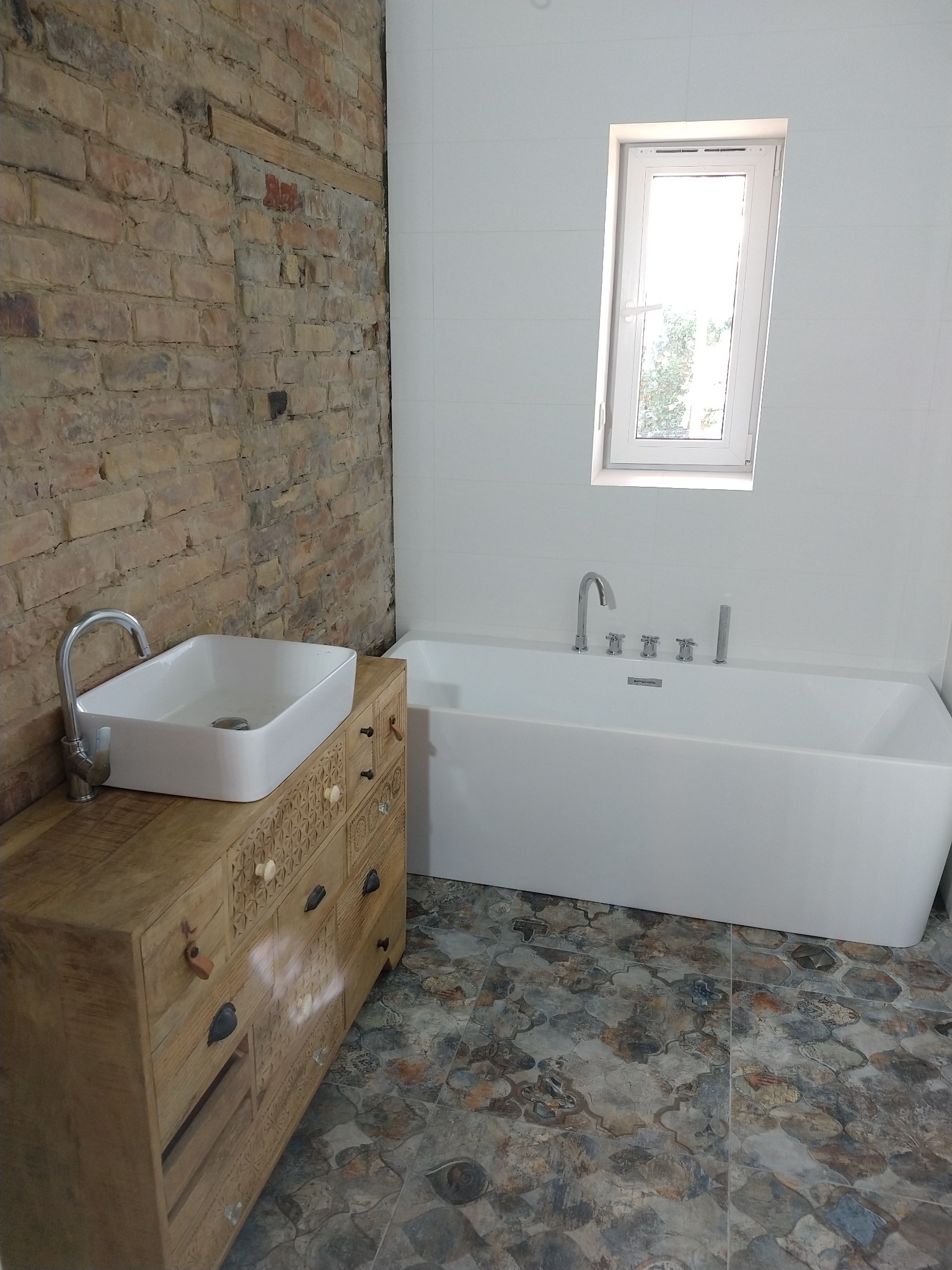 Nowoczesna łazienka z wanną prostokątną, umywalką nablatową na zdobionej komodzie z jasnego drewna, ścianą z cegły i podłogą z wzorzystych płytek ceramicznych.