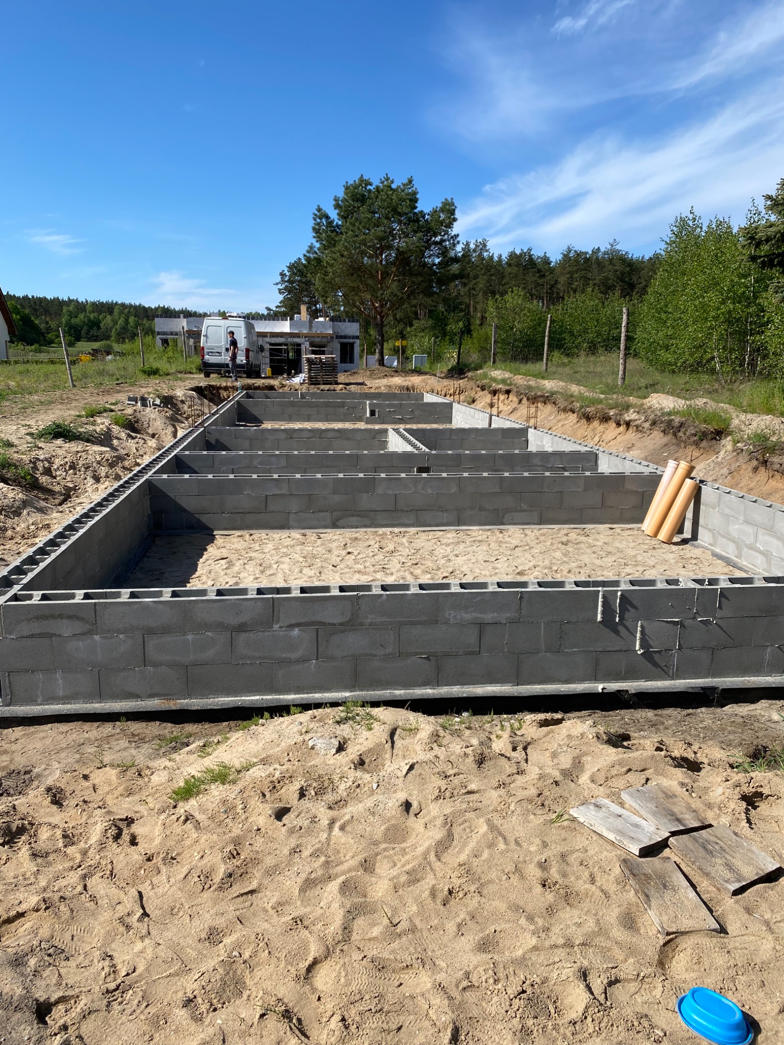 Fundamenty domu jednorodzinnego w trakcie budowy, bloczki betonowe ułożone w wykopie wypełnionym piaskiem, widoczne rury kanalizacyjne i zaparkowany bus budowlany w tle.