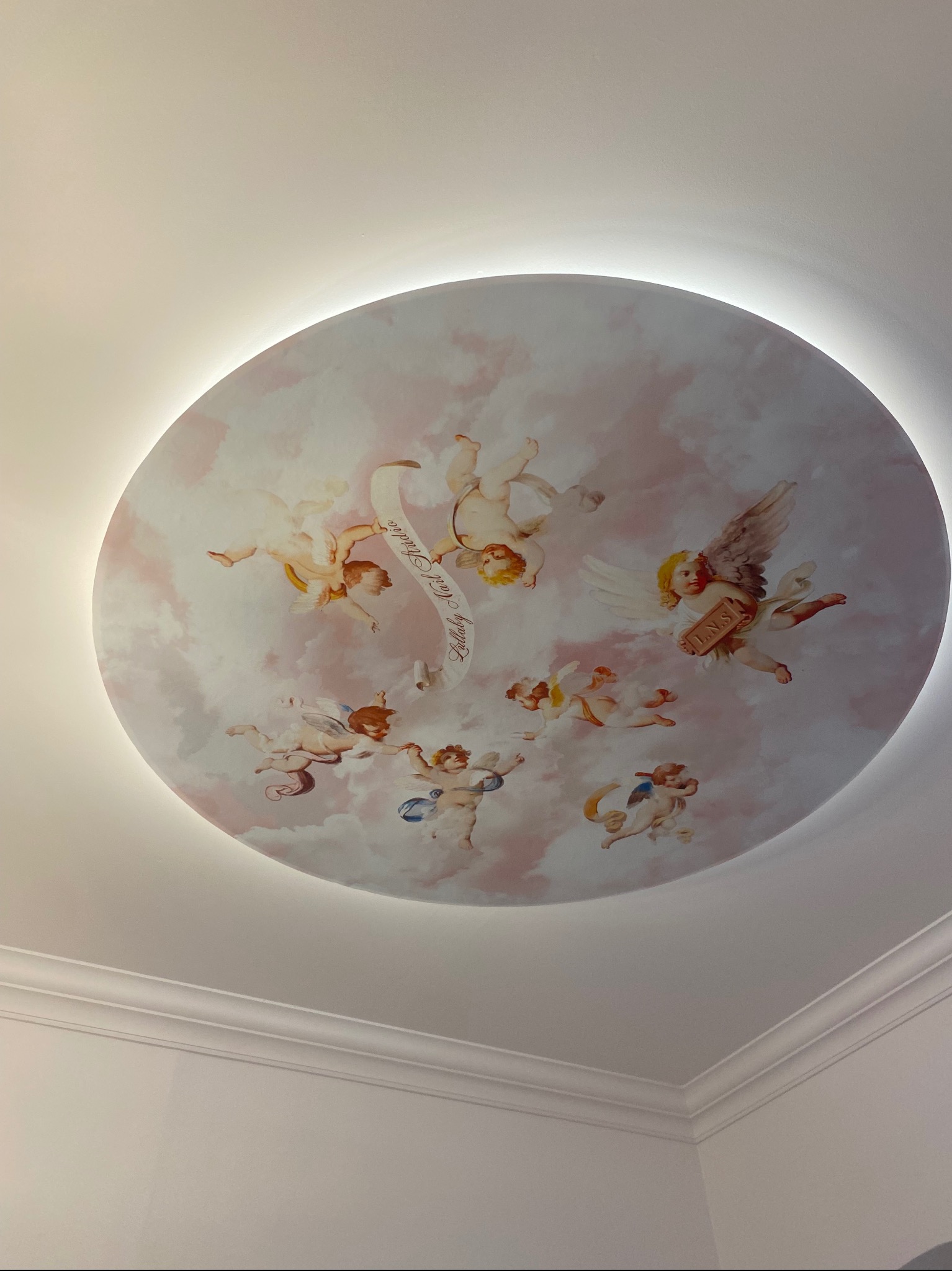 Okrągła dekoracja sufitowa z motywem aniołków na tle chmur, podświetlona od tyłu, widoczna z dołu, z detalami sztukaterii na styku ze ścianą.