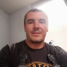 Portret mężczyzny w szarym kombinezonie roboczym z butelką wody za plecami, stojącego w jasnym, odnowionym pomieszczeniu.