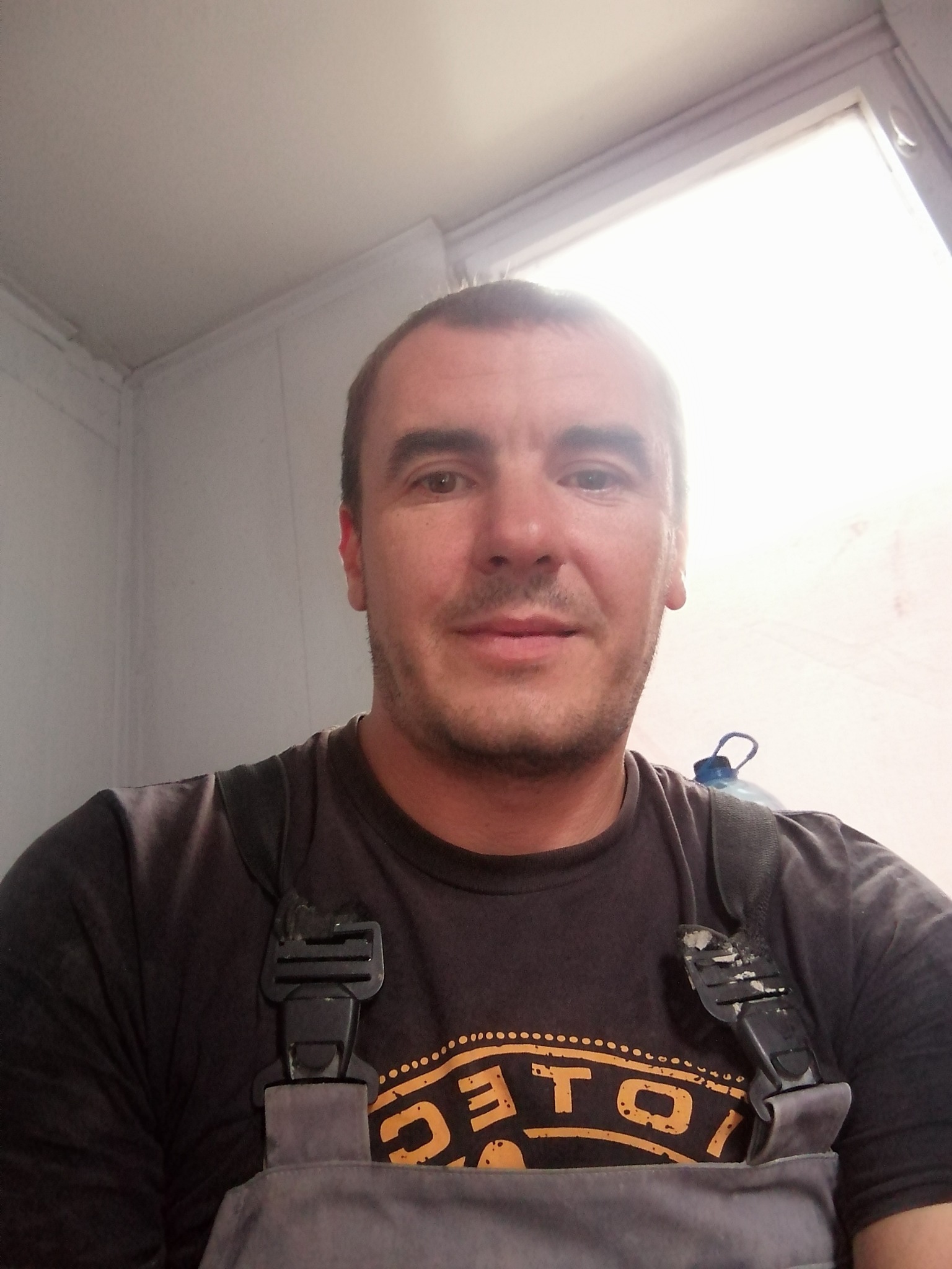 Portret mężczyzny w szarym kombinezonie roboczym z butelką wody za plecami, stojącego w jasnym, odnowionym pomieszczeniu.