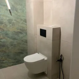 Nowoczesna łazienka z podwieszaną toaletą, beżowymi płytkami i zielonym akcentem marmurowej ściany, widoczny bidetta w kolorze czarnym.