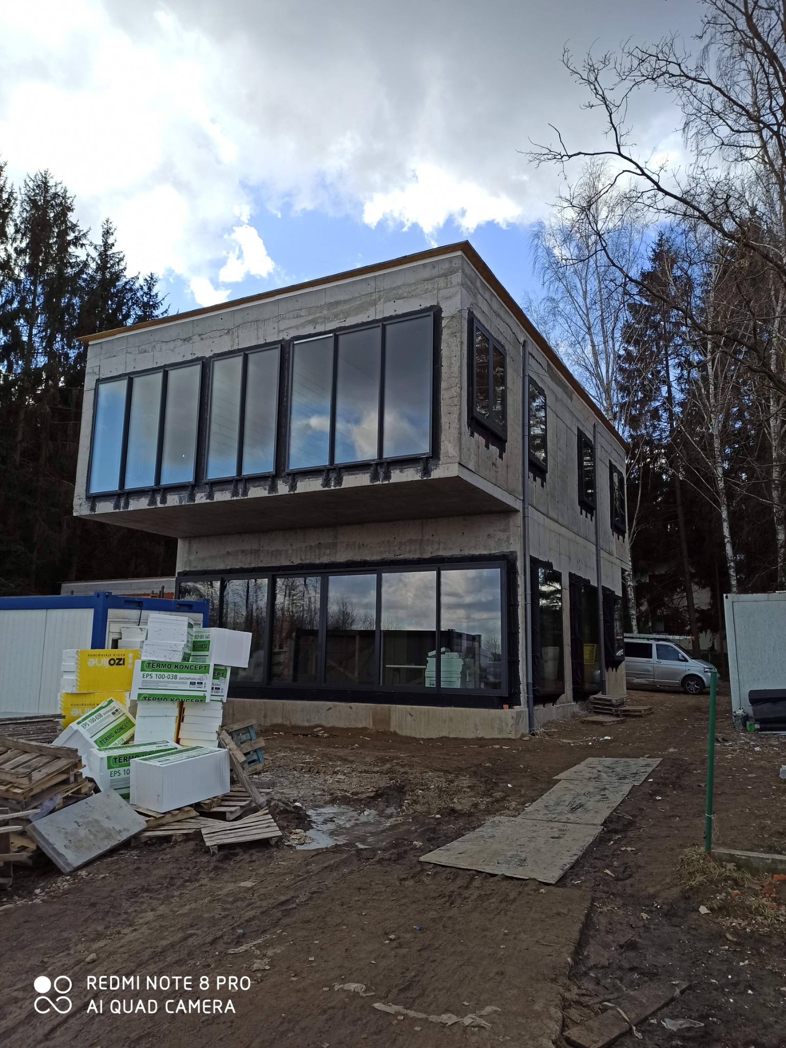 Surowy stan domu murowanego z betonu komórkowego i dużymi oknami z czarnymi ramami, widoczne ocieplenie styropianowe na paletach, teren budowy z błotem, w tle drzewa i pochmurne niebo.