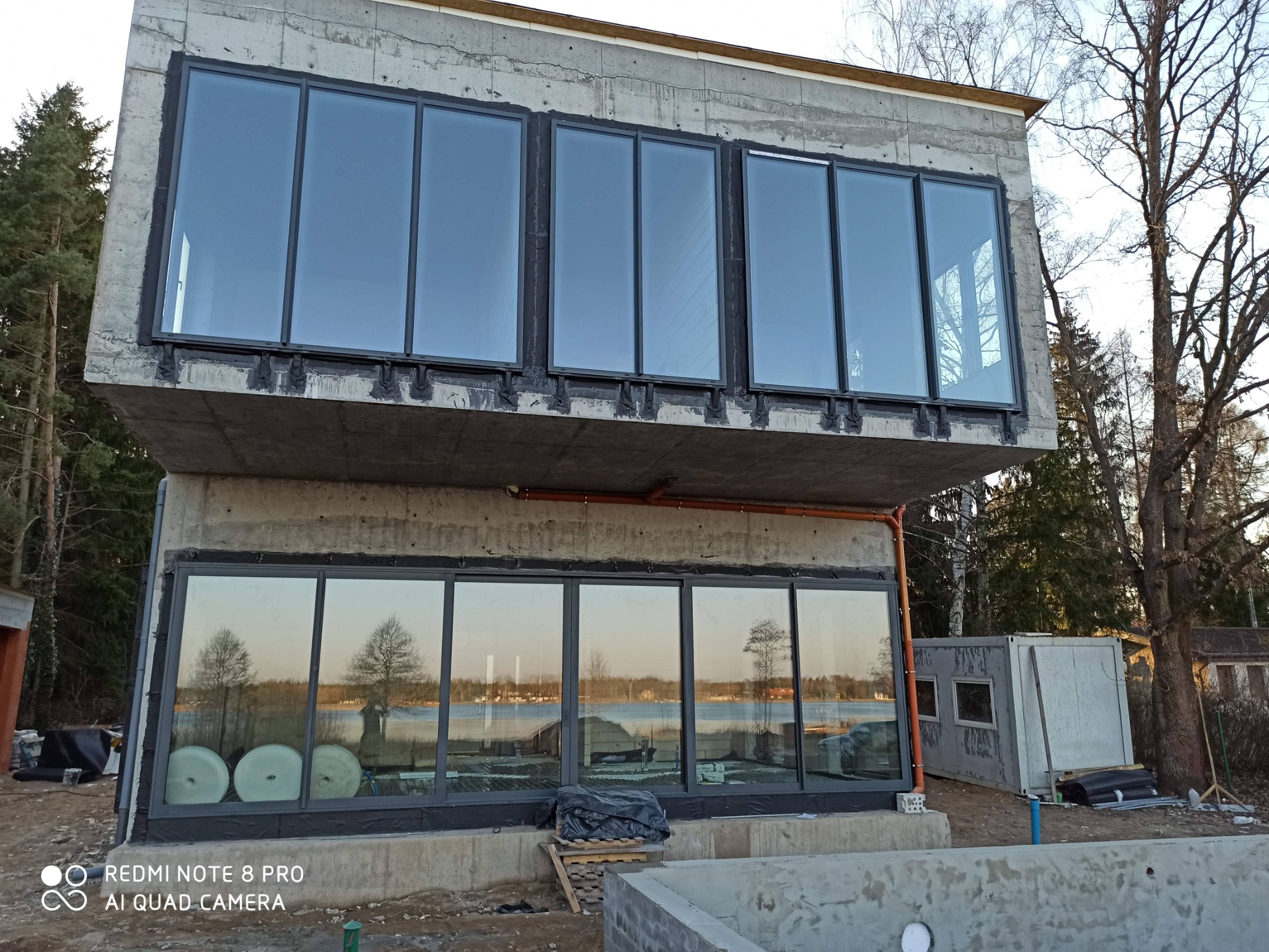 Surowy, dwukondygnacyjny dom z betonu z dużymi oknami, widoczne elementy konstrukcyjne i instalacje, otoczenie w stanie budowy.