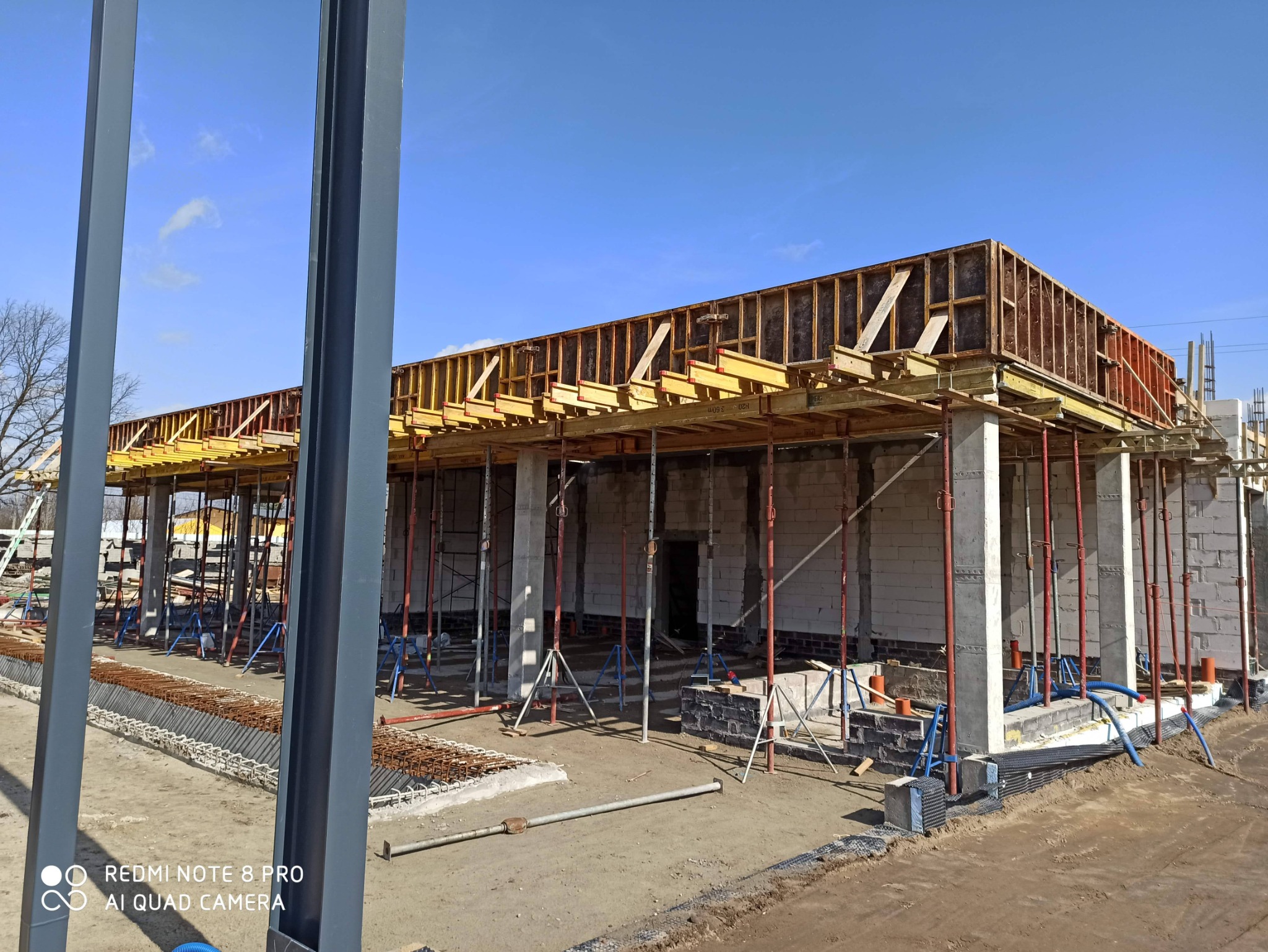 Budowa budynku z białych bloczków, widoczne szalunki i podpory, niebieskie rury wystające z ziemi.