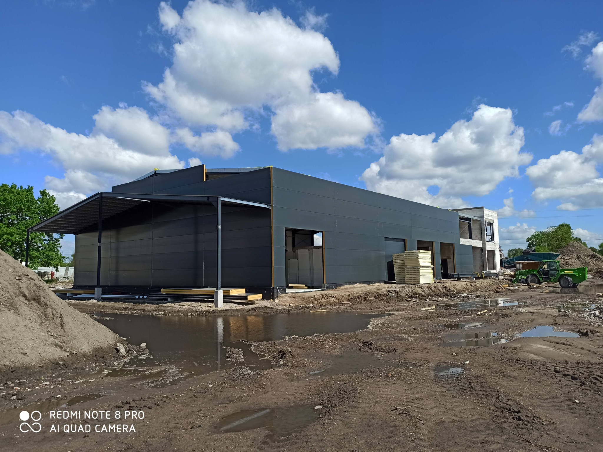 Budowa hali przemysłowej z paneli warstwowych w trakcie realizacji, widoczne elementy konstrukcyjne, stos paneli oraz teren budowy z kałużami po deszczu, na dalszym planie widać budynek w stanie...