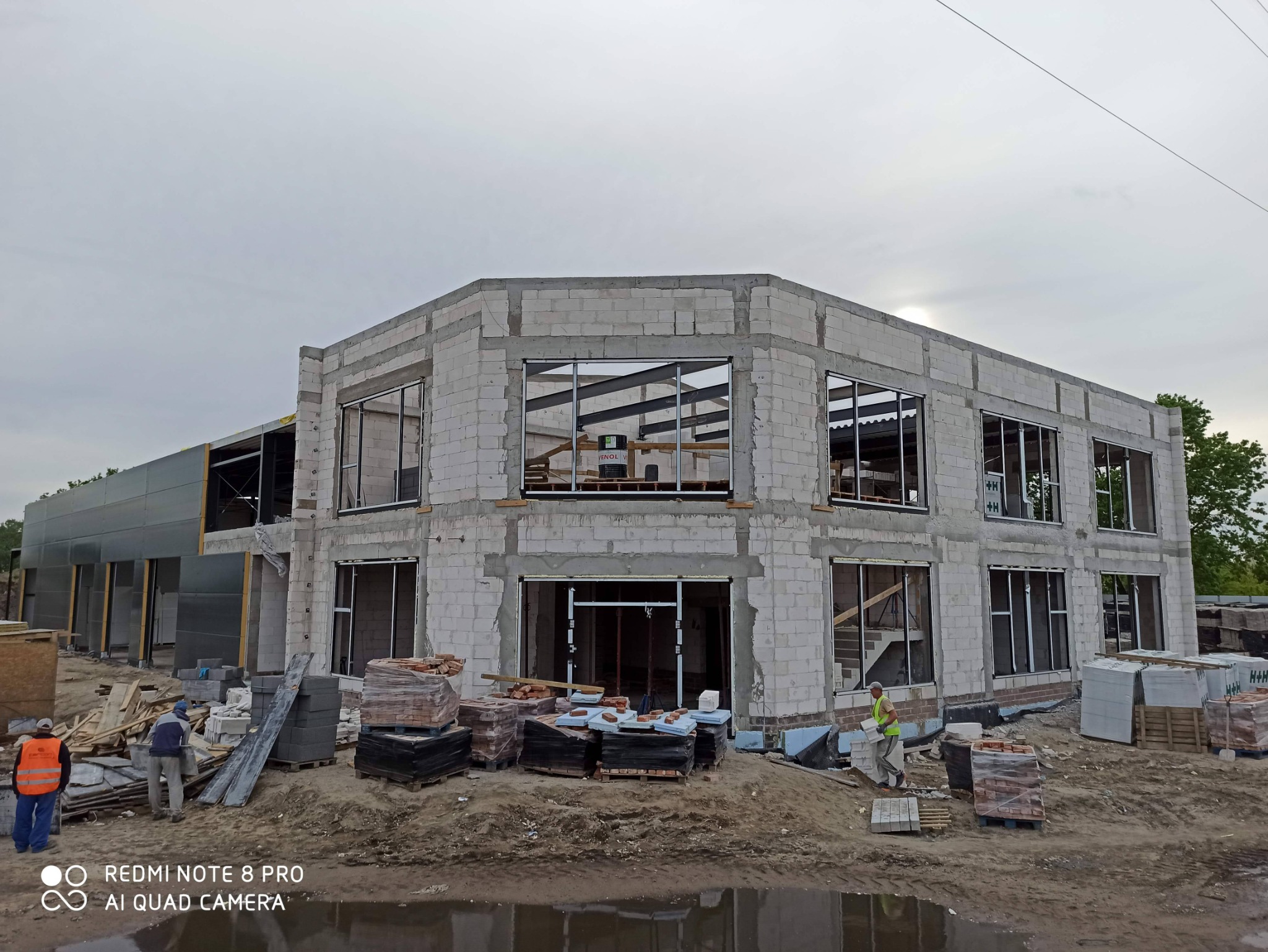Budowa dwukondygnacyjnego budynku z bloczków, widoczne stalowe ramy okienne, palety z cegłami i pracownicy w odblaskowych kamizelkach, pochmurne niebo.