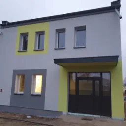 Dwukondygnacyjny budynek z elewacją w kolorach szarości i jasnożółtym, z widocznymi oknami i drzwiami wejściowymi, na tle pochmurnego nieba i placu budowy.