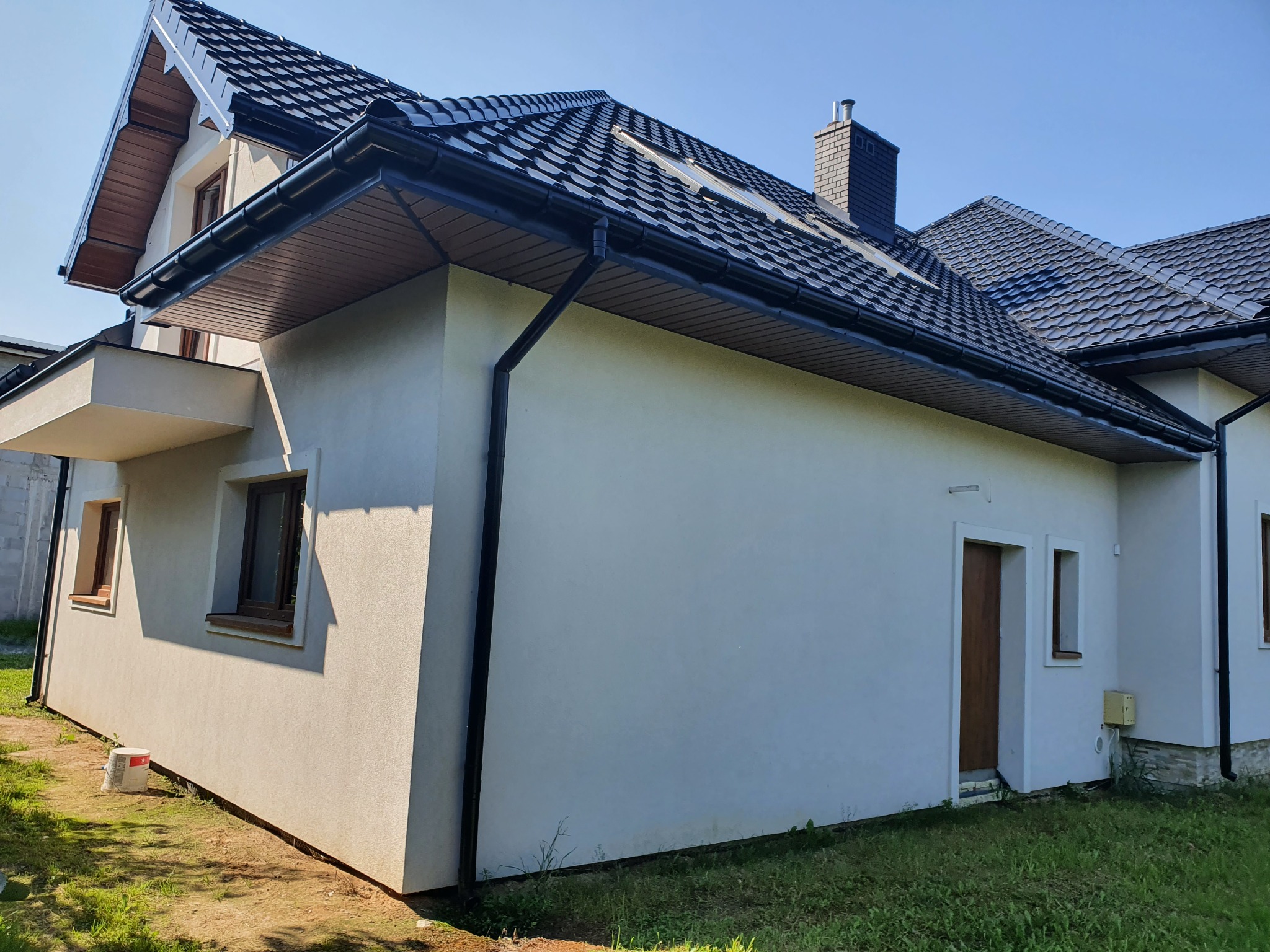 Świeżo otynkowana elewacja domu jednorodzinnego z grafitową dachówką, widoczne rynny i fragment trawnika, w tle błękitne niebo.