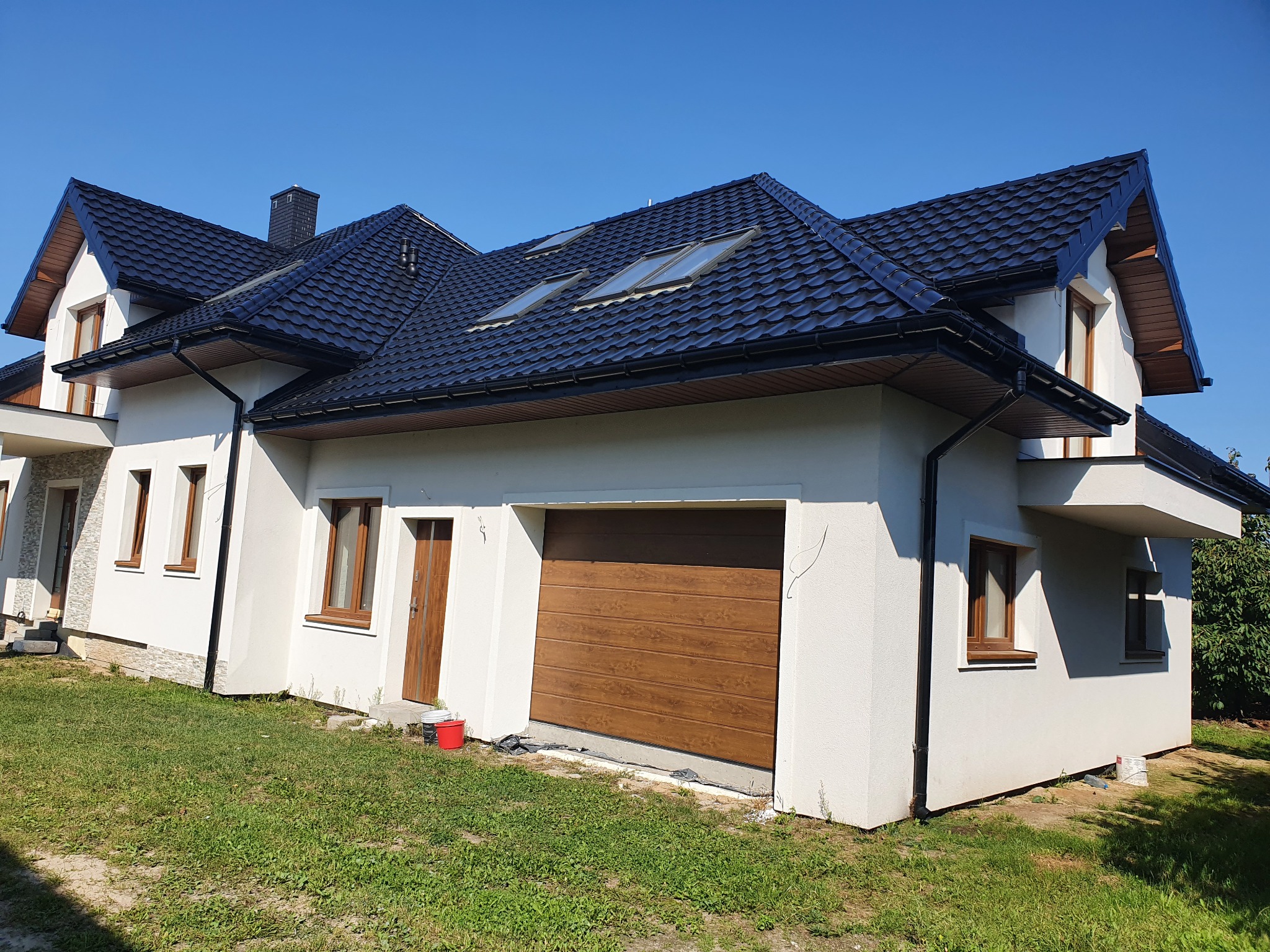 Nowoczesny dom jednorodzinny z ciemną dachówką, jasną elewacją i drewnianą bramą garażową, uchwycony w słoneczny dzień z perspektywy trawnika, widoczne ślady po pracach budowlanych.