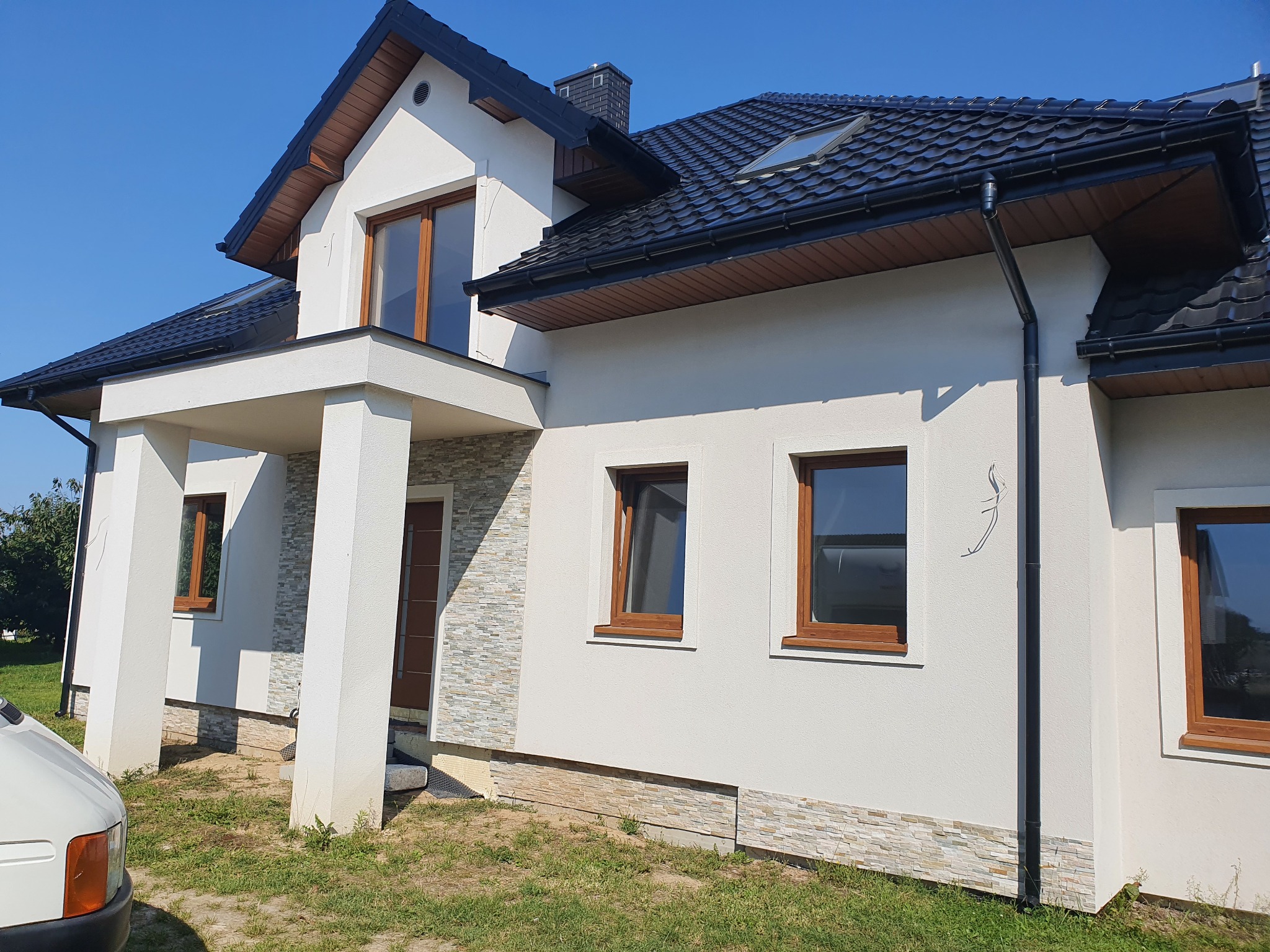 Nowoczesny dom jednorodzinny z białą elewacją, brązowymi oknami i ciemną dachówką, z fragmentem samochodu dostawczego widocznym w kadrze.