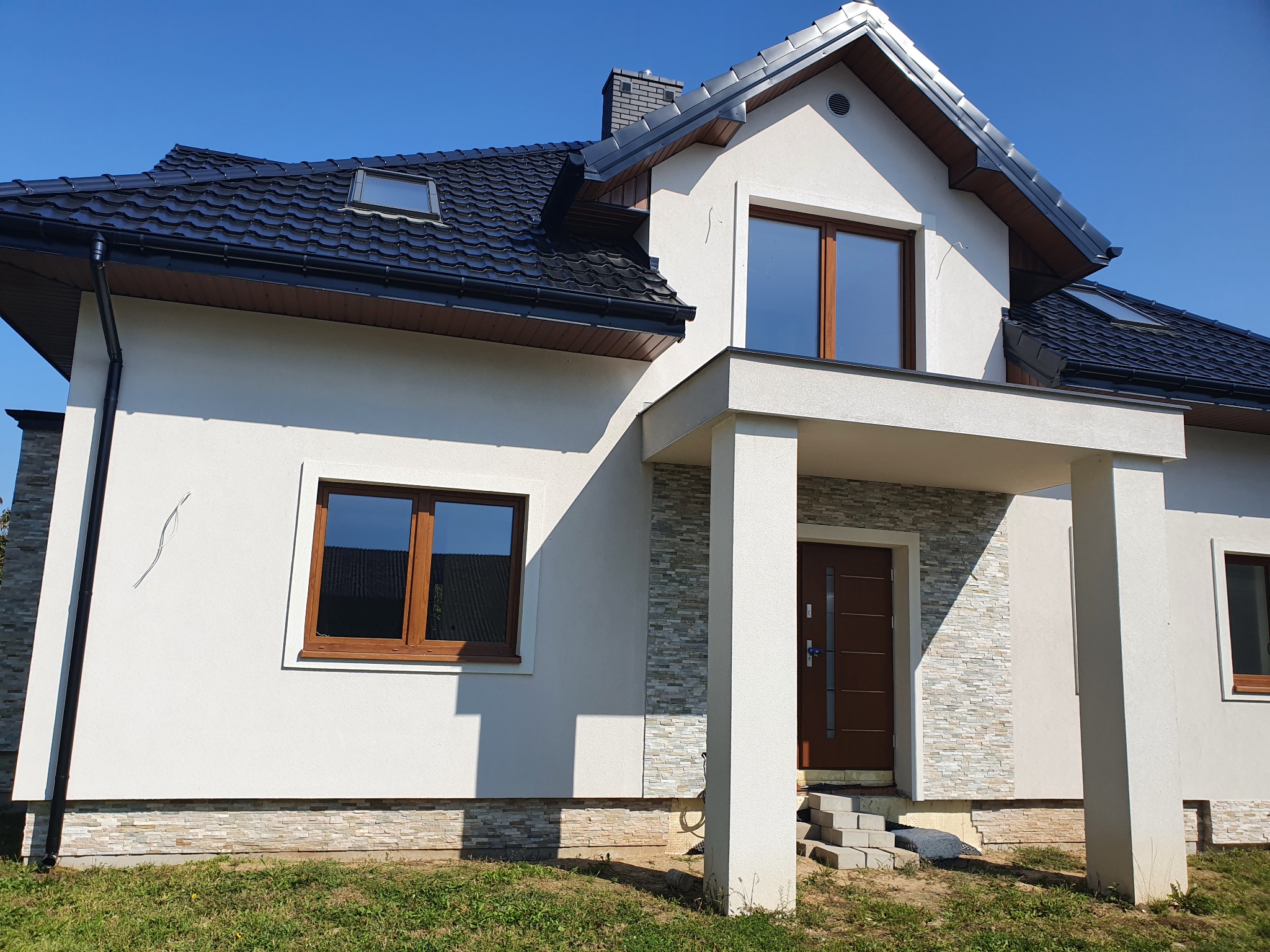 Nowoczesny dom jednorodzinny z białą elewacją, ciemną dachówką i drewnianymi oknami, widok z zewnątrz na fragment fasady z wejściem, dekoracyjna okładzina kamienna na fragmentach ścian.