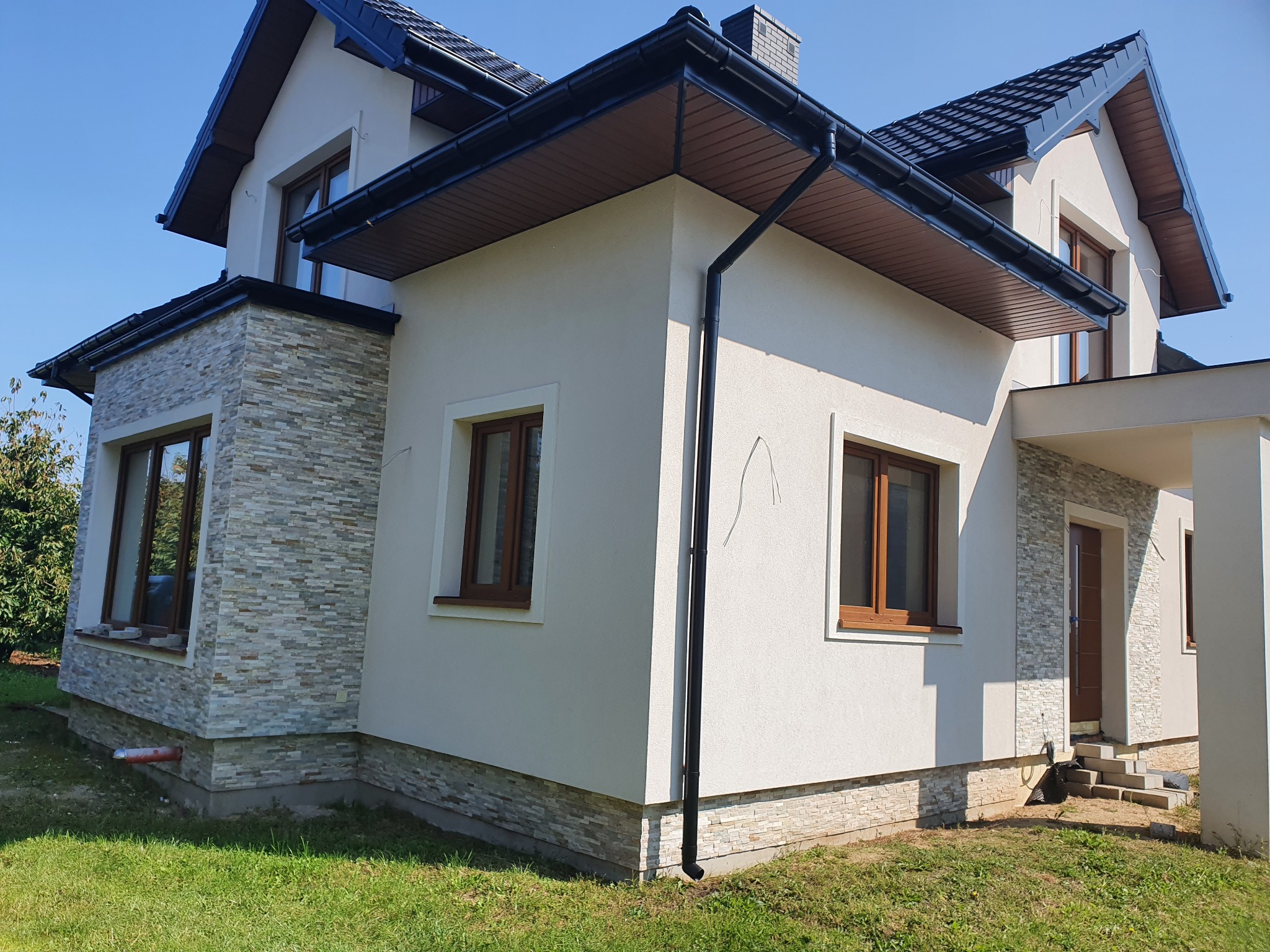 Nowoczesny dom jednorodzinny z elewacją w odcieniach bieli i szarości, z elementami kamiennymi, ciemną dachówką i brązowymi oknami, w słoneczny dzień.