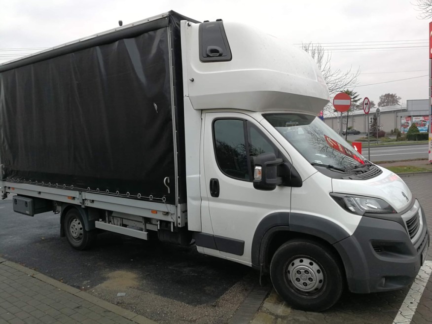 Biały samochód dostawczy z czarną plandeką na parkingu, widoczny znak drogowy w tle.