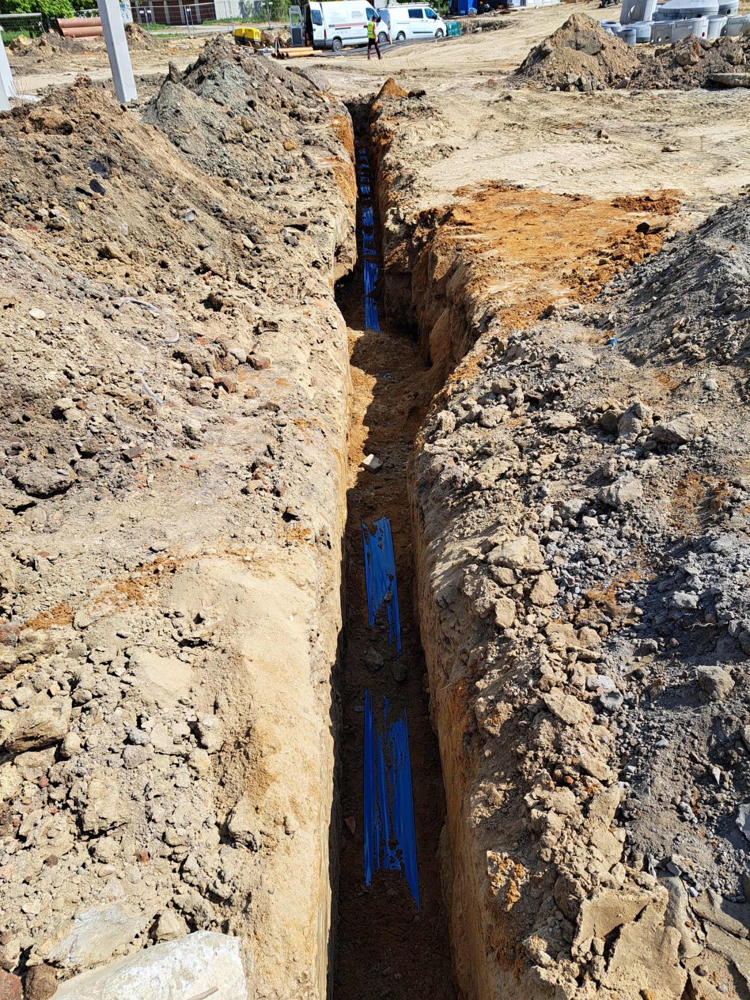 Wykop z ułożonymi niebieskimi rurami wodociągowymi lub kanalizacyjnymi, widok z góry, w tle widoczny samochód dostawczy i elementy budowy.