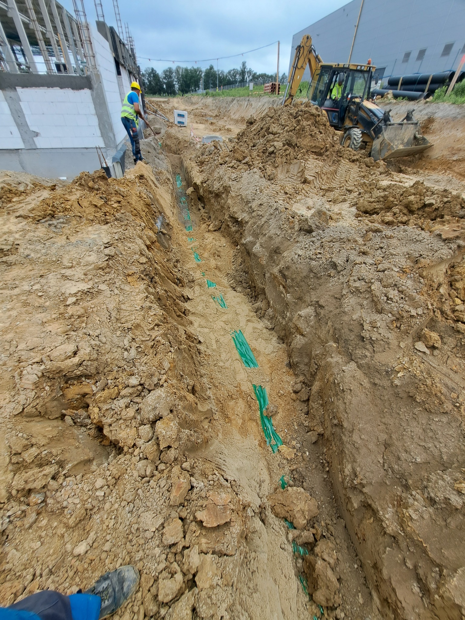 Wykop z ułożonym rurociągiem w zielonej otulinie, pracownik z łopatą i koparko-ładowarka w tle, budynek w stanie surowym w oddali.
