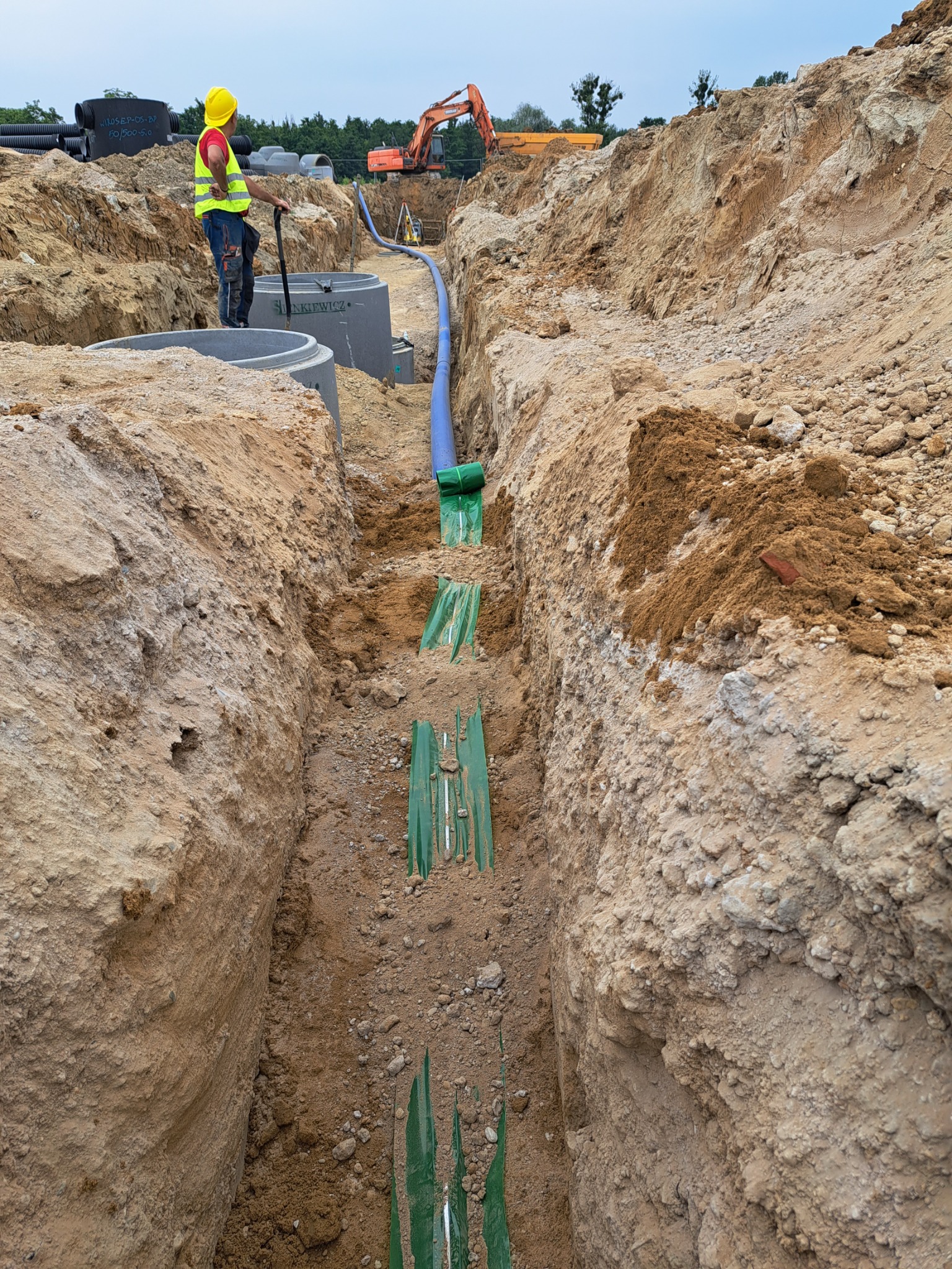 Głęboki wykop z ułożonym rurociągiem, widoczny pracownik w żółtym kasku i kamizelce odblaskowej oparty o szpadel, betonowe studzienki i koparka w tle.