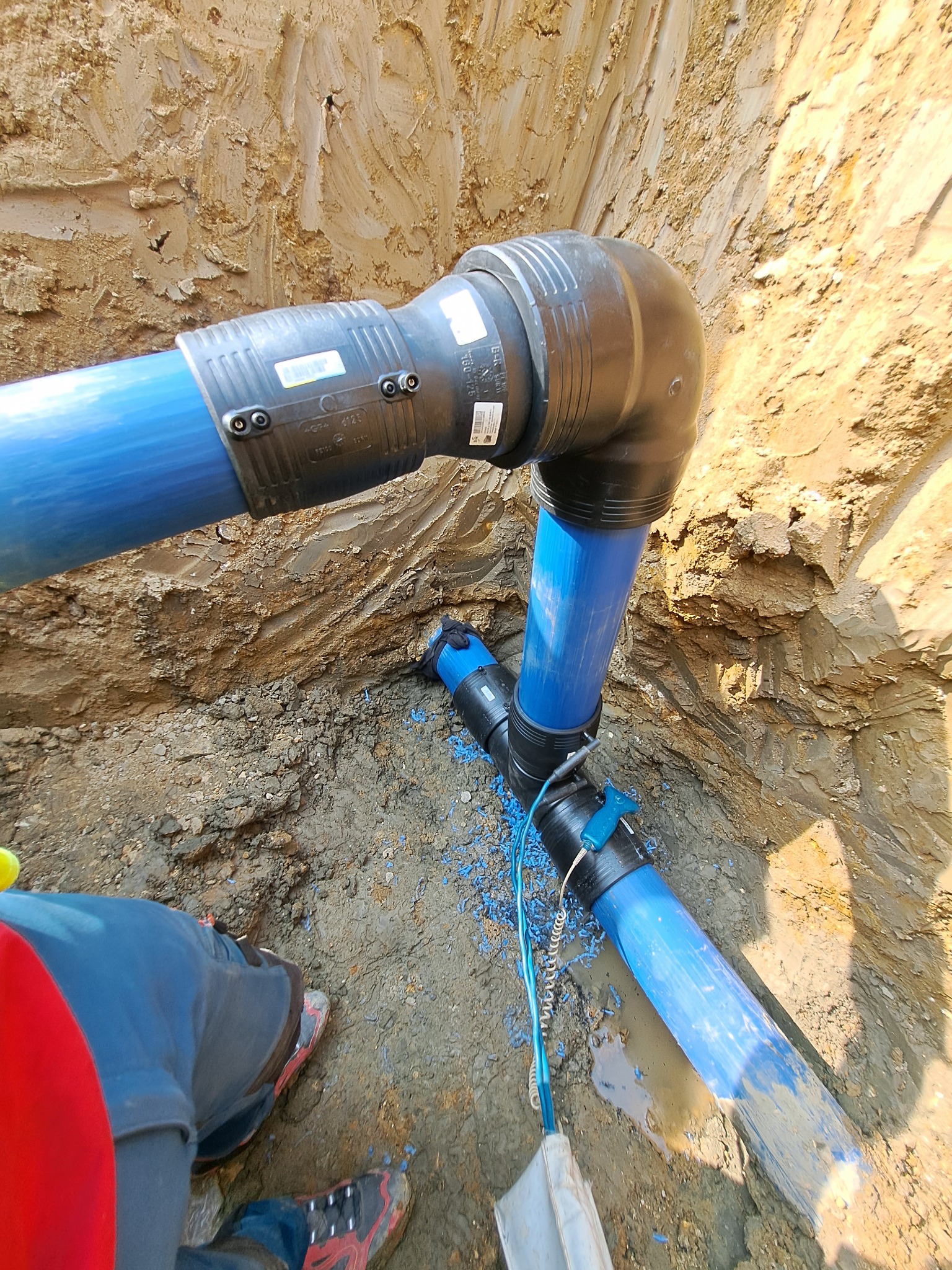 Niebieskie rury wodociągowe z czarnymi złączkami zakopane w wykopie ziemnym, widok z góry, fragment odzieży roboczej w lewym dolnym rogu.