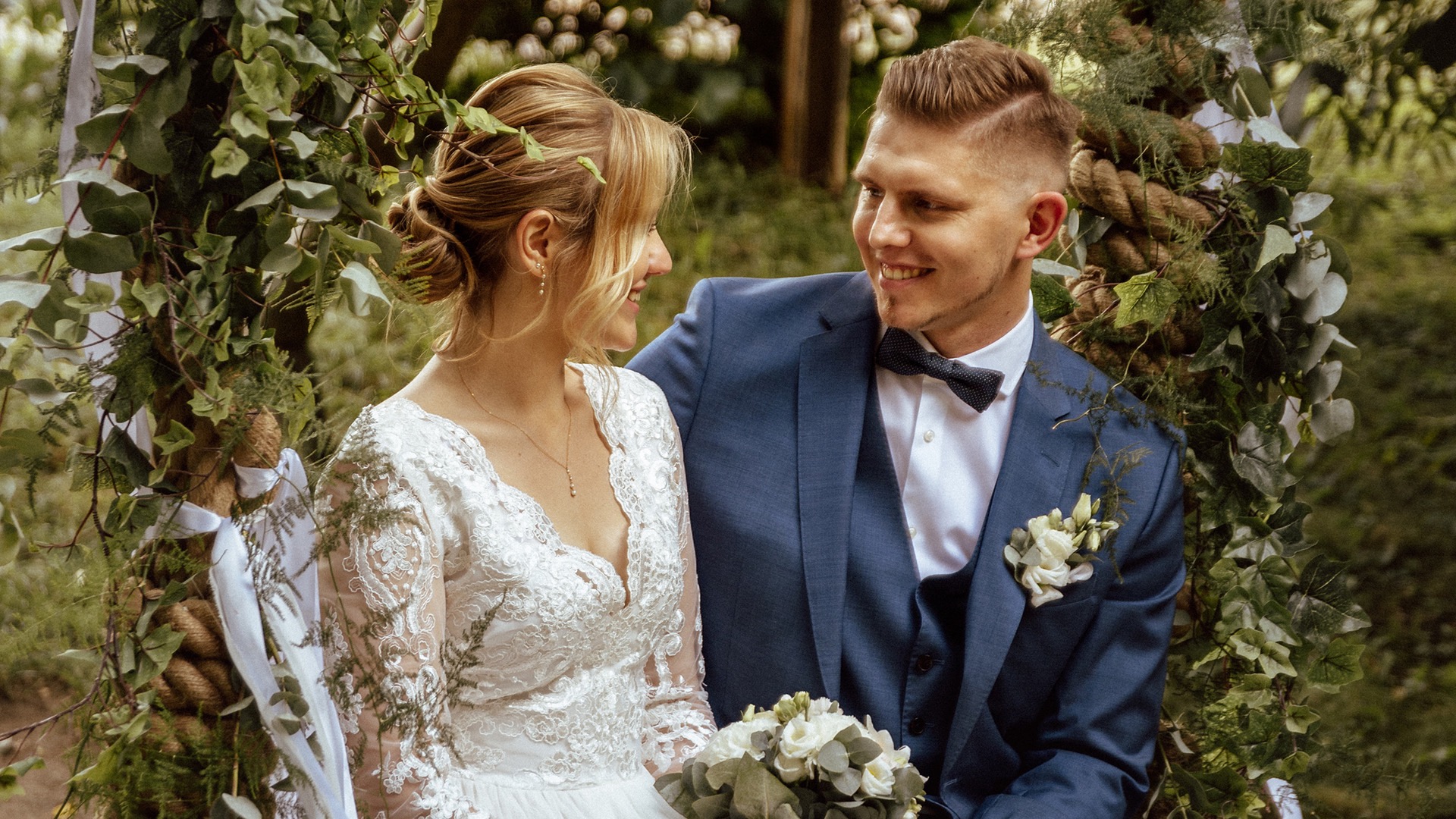 Uśmiechnięta para młoda, panna młoda w koronkowej sukni z bukietem, pan młody w niebieskim garniturze z muszką i butonierką, siedząca na huśtawce ozdobionej zielenią, wymieniająca spojrzenia.