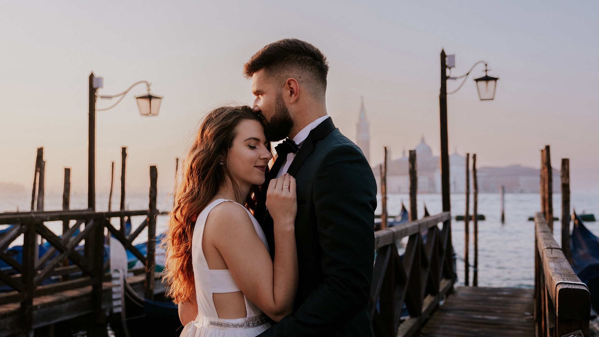 Para młoda w Wenecji podczas zachodu słońca, mężczyzna całuje kobietę w czoło na drewnianym pomoście z gondolami w tle.