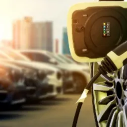 Ładowanie samochodu elektrycznego z widocznym podświetlanym na zielono gniazdem ładowania, na tle rzędu zaparkowanych aut i budynków miejskich w oddali.