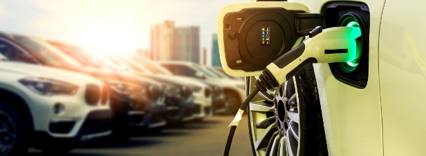 Ładowanie samochodu elektrycznego z widocznym podświetlanym na zielono gniazdem ładowania, na tle rzędu zaparkowanych aut i budynków miejskich w oddali.