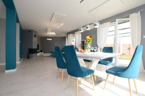 Nowoczesne wnętrze jadalni z białym stołem, turkusowymi krzesłami i płaskimi panelami grzewczymi na suficie.