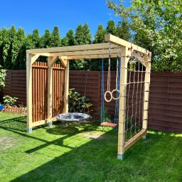 Abilmont - Drewniana konstrukcja placu zabaw w ogrodzie z huśtawkami, drabinką sznurową i ścianką z pionowych listew, otoczona zielenią i brązowym płotem.