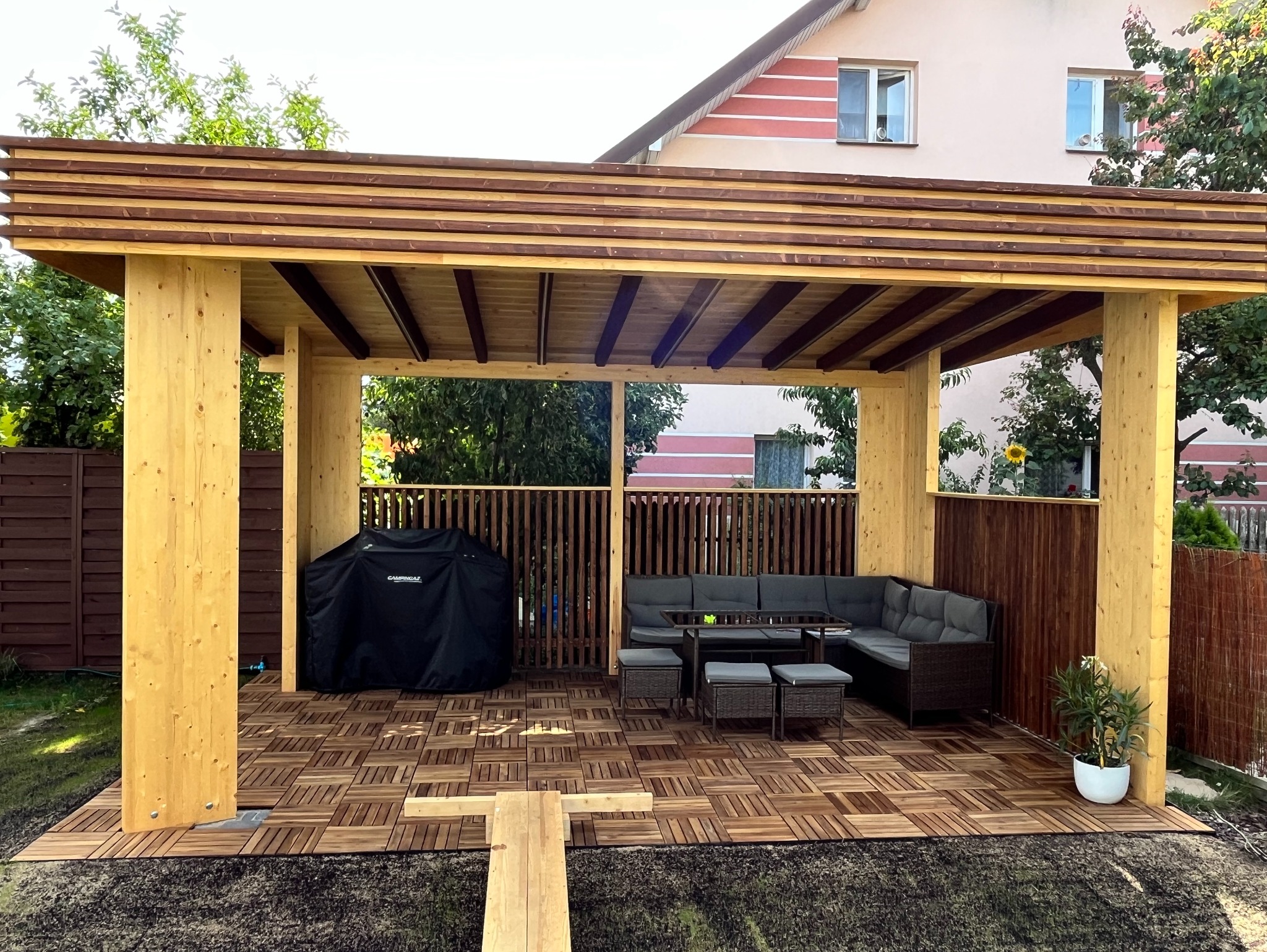 Drewniana altana z meblami ogrodowymi, grillem pod pokrowcem i podłogą z płytek, na tle domu jednorodzinnego i ogrodzenia.